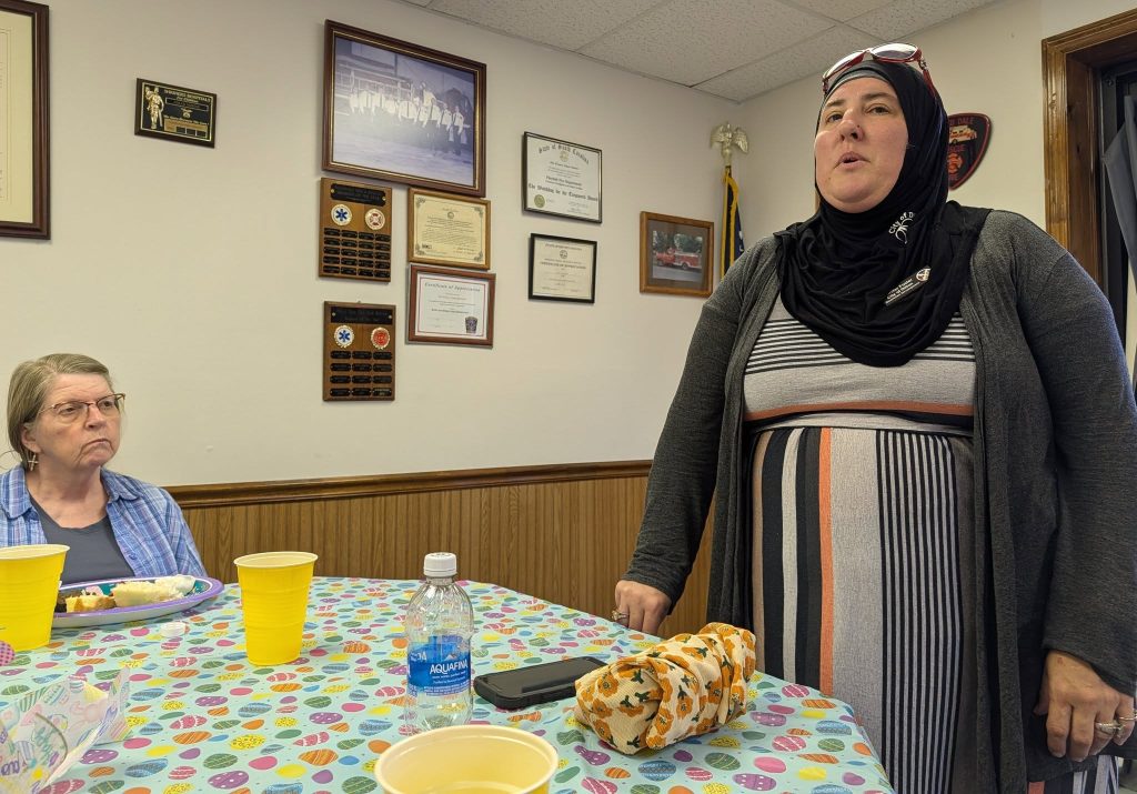 Two women in a room with certificates and plaques on the wall, sitting and standing by a table with food, drinks, and a colorful tablecloth.