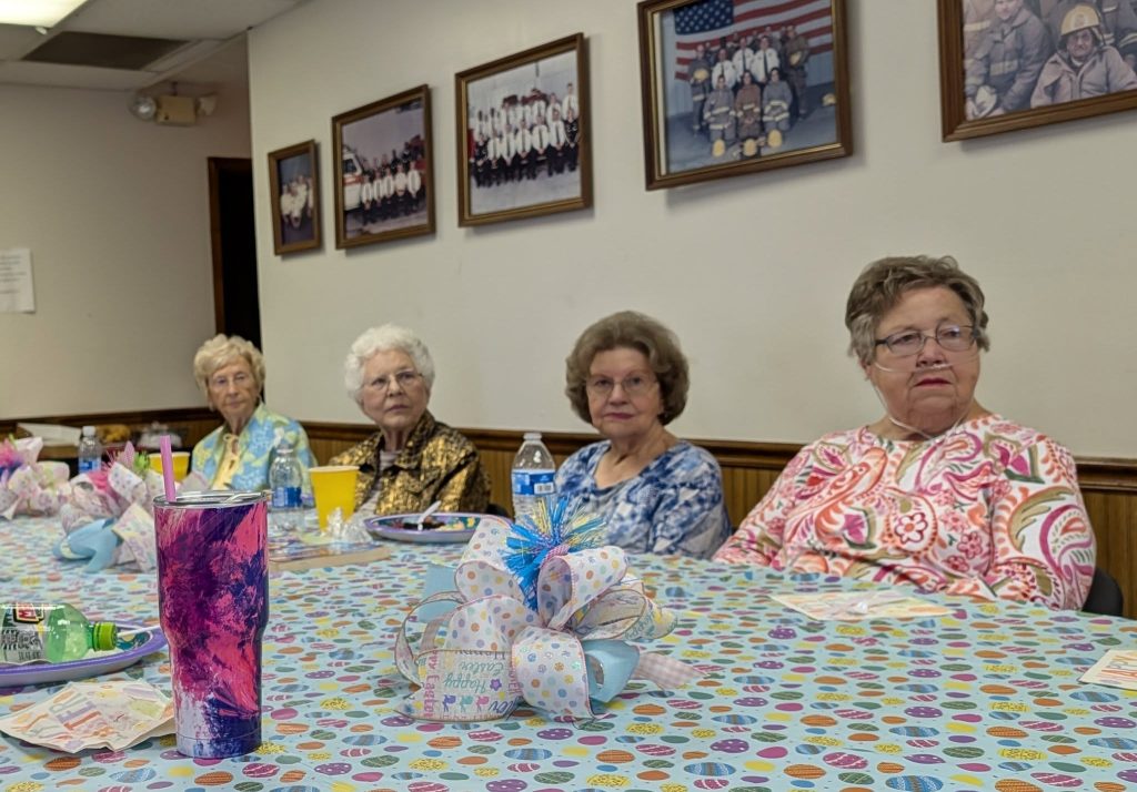 Four elderly women sit at a decorated table in a community room, with colorful drinks and Easter-themed tablecloth, and framed group photos hanging on the wall behind them.