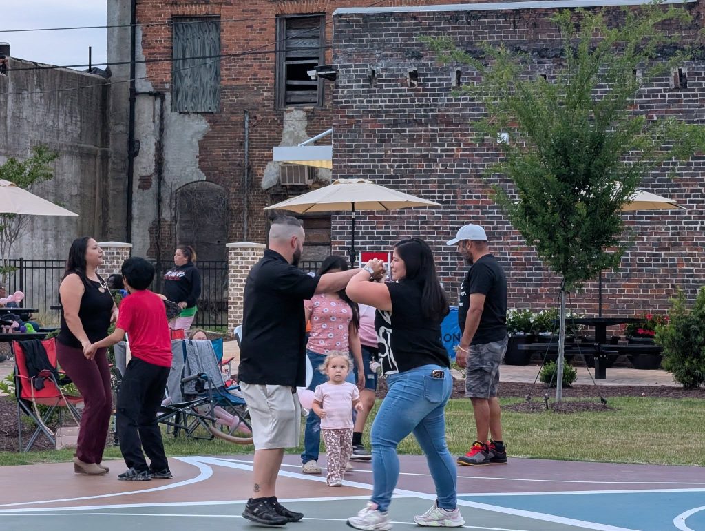 People dancing and socializing outdoors on a basketball court, with a brick building and umbrellas in the background. A small child stands in the foreground.