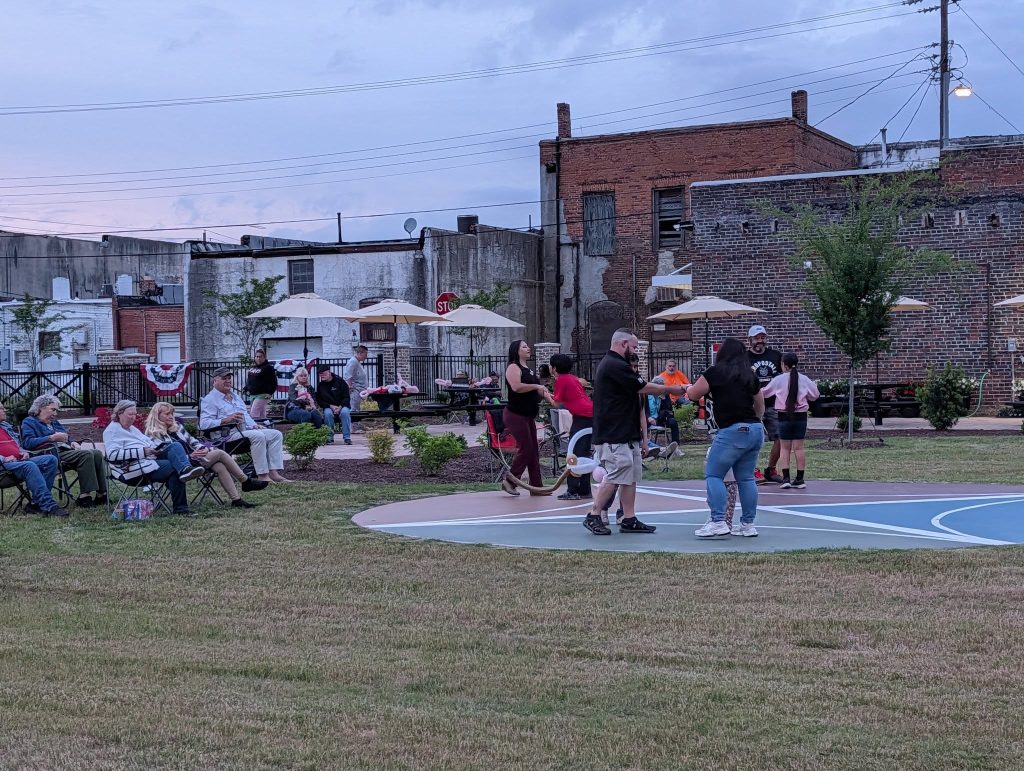 People dancing on a basketball court while others sit and watch in a park, with brick buildings and umbrellas in the background.