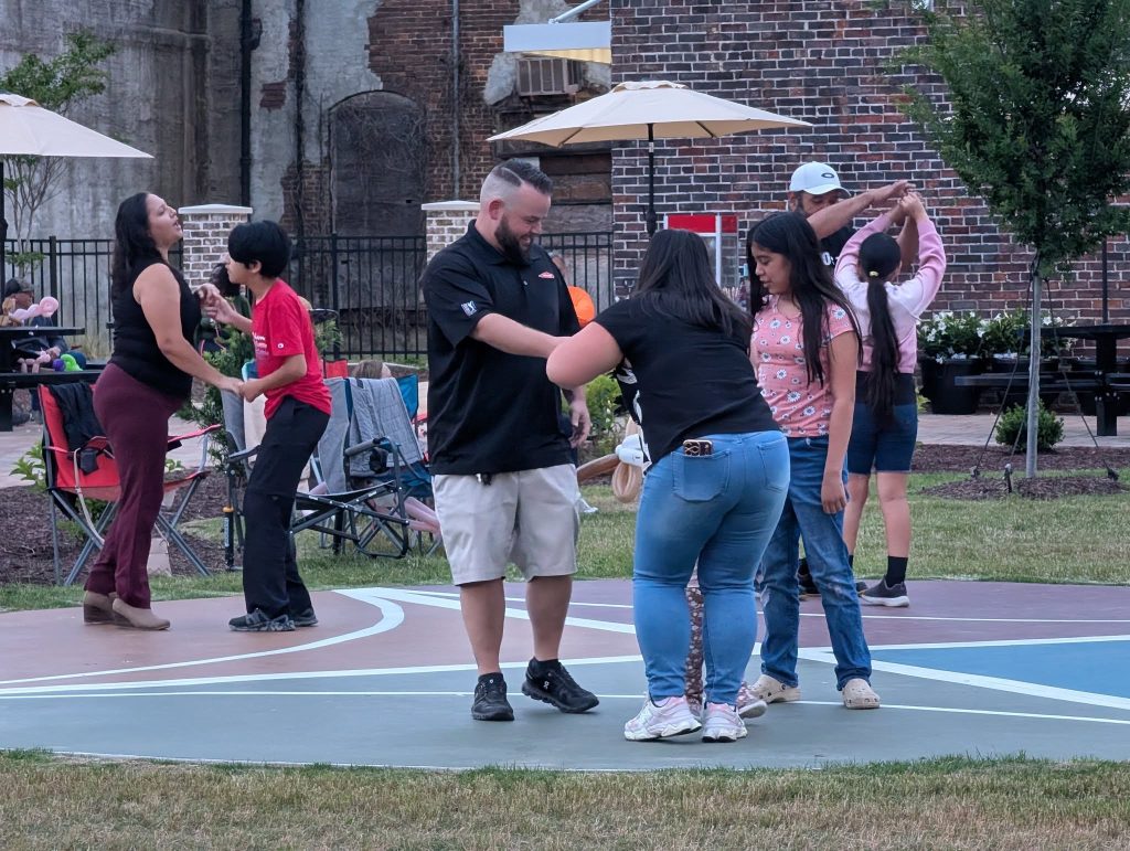 Groups of people dancing together outdoors on a court, surrounded by grass, umbrellas, and a brick building in the background.