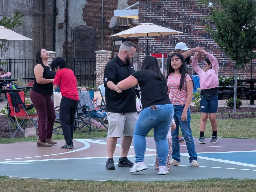 People of different ages dancing and playing together outside on a court, with chairs and umbrellas set up in the background near a brick wall.