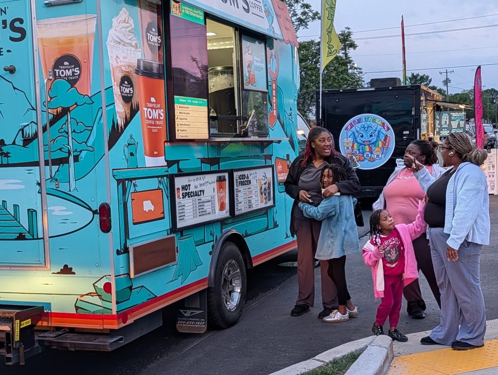 A group of women and children stand near a colorful coffee truck, talking and smiling at an outdoor event with other food trucks in the background.