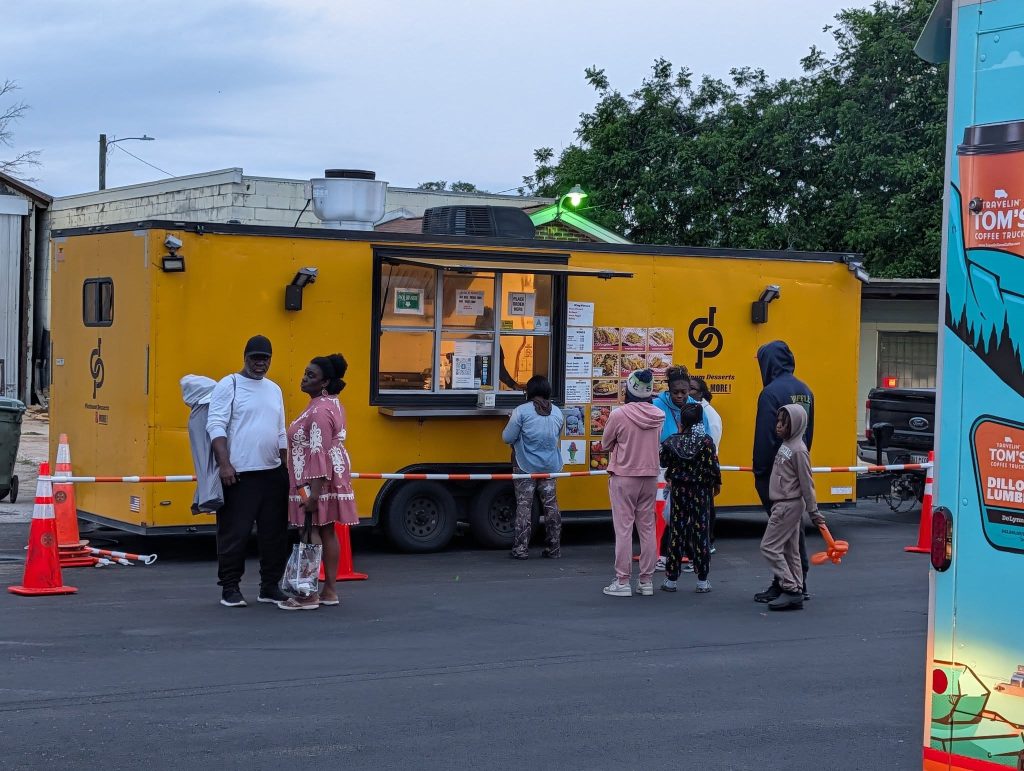 People standing in line at a yellow food truck on a street, with menus displayed and orange cones nearby, during the evening.