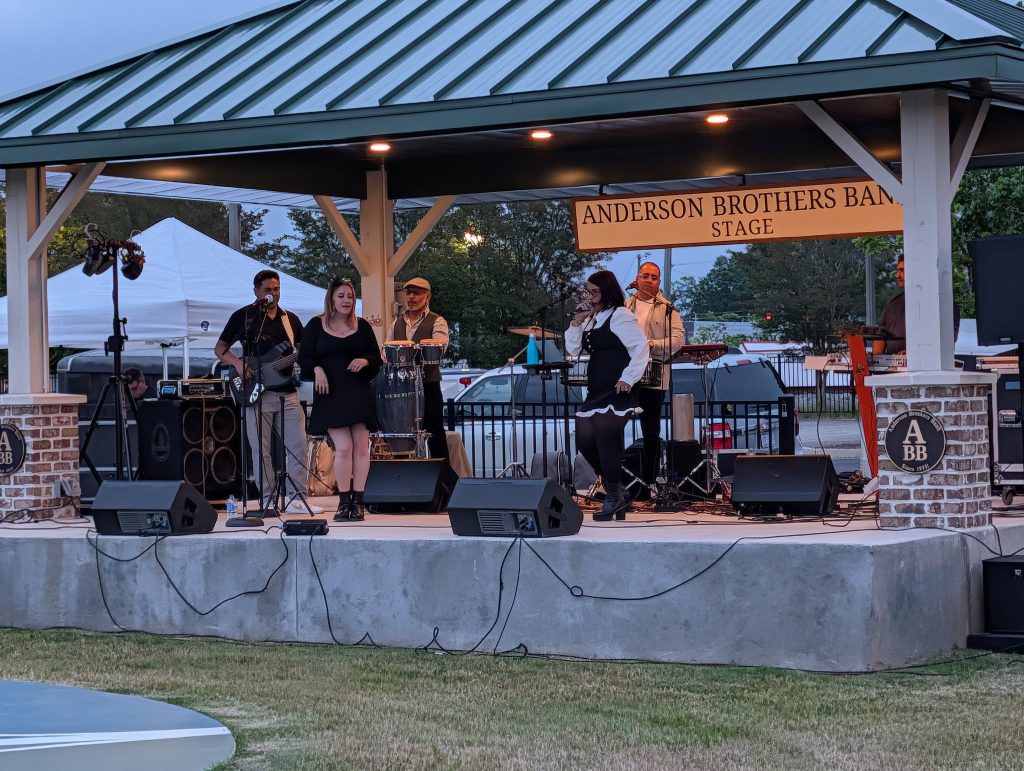 A band performs on an outdoor stage labeled "Anderson Brothers Band Stage," with musicians playing instruments and two singers under a green metal roof.