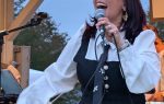 A woman with red glasses sings passionately into a microphone on an outdoor stage, wearing a black and white outfit with a decorative tie.