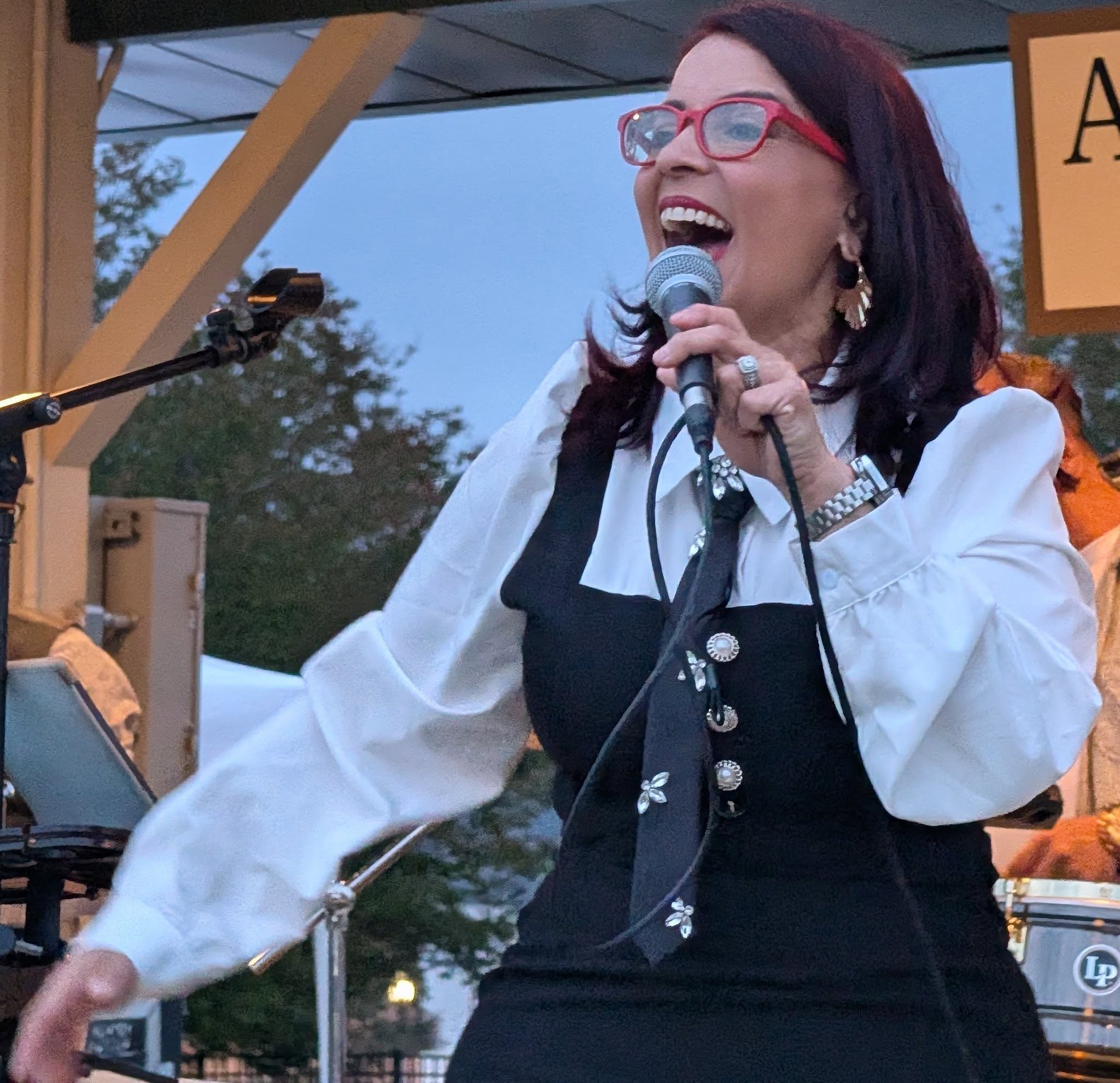 A woman with red glasses sings passionately into a microphone on an outdoor stage, wearing a black and white outfit with a decorative tie.