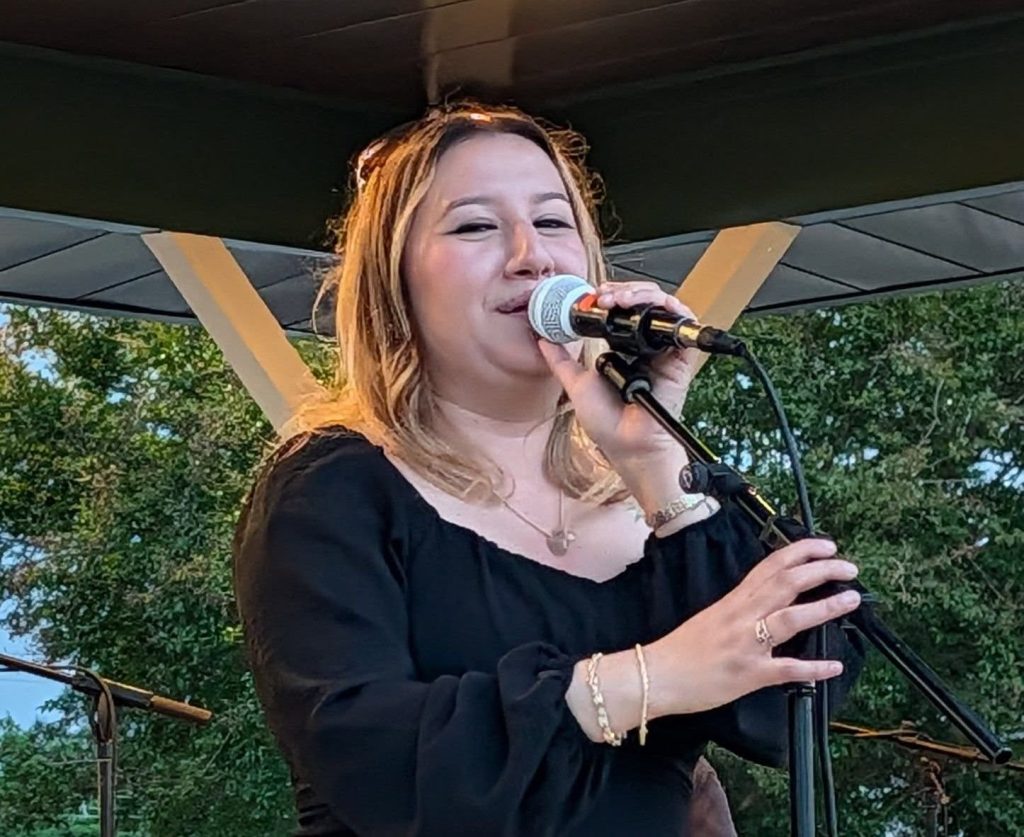 A woman in a black dress sings into a microphone on an outdoor stage, with green trees and metal beams in the background.