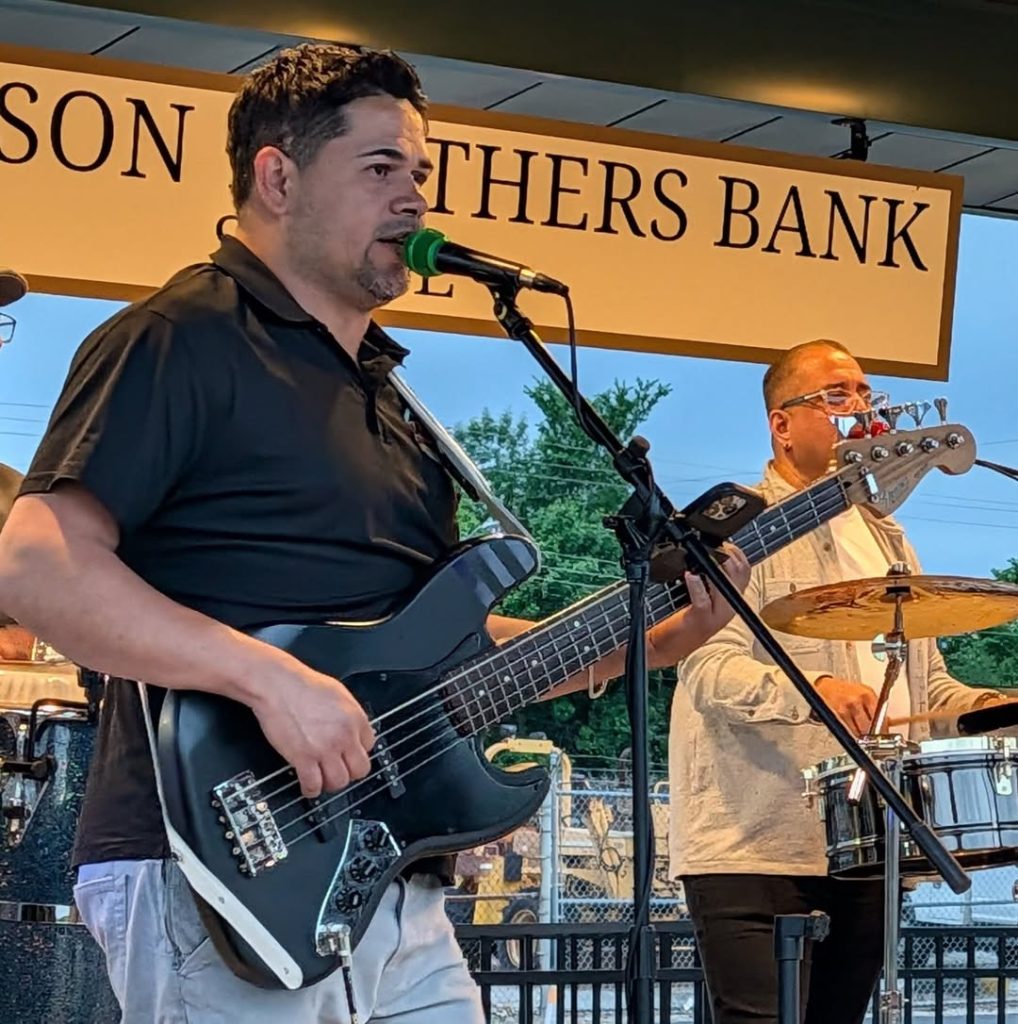A man plays electric bass guitar and sings into a microphone on stage, with another musician playing drums in the background.