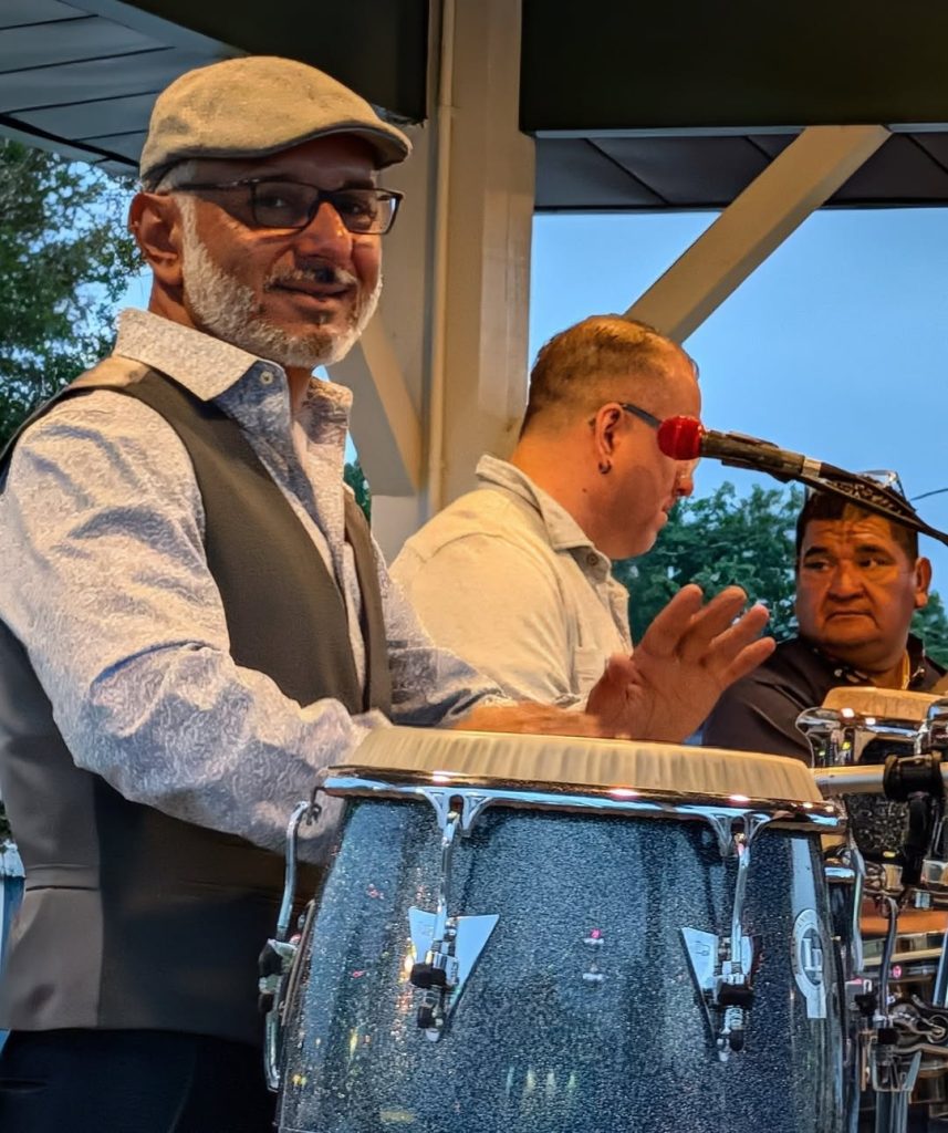 Three men performing music outdoors; one is playing conga drums, wearing glasses, a cap, and a vest, while the others focus on microphones and percussion instruments.