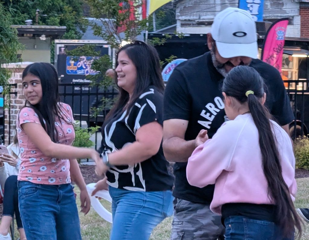 Four people, including two adults and two children, are dancing and smiling together outdoors in a casual, festive environment during the daytime.
