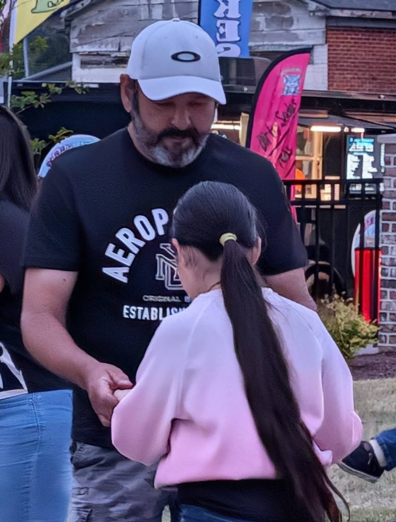 A man wearing a white cap holds hands with a girl in a pink sweatshirt at an outdoor event with colorful flags in the background.