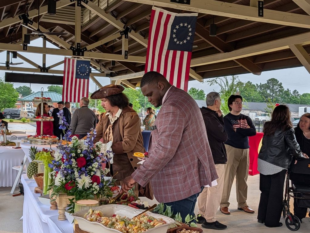People gather at a covered outdoor event with food tables, colonial-era costumes, and American flags with 13 stars hanging from the ceiling.