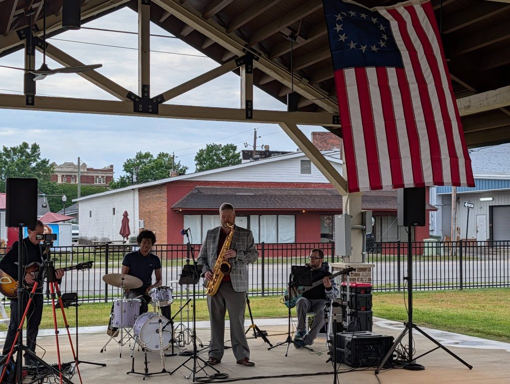 A jazz band plays under a pavilion with drums, saxophone, guitar, and bass. An American flag hangs above. Buildings are visible in the background.