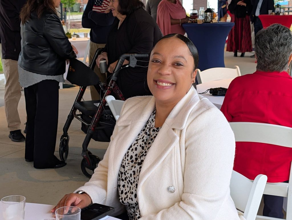 Smiling woman in a white coat sits at a table during an outdoor social event, with people mingling and talking in the background.