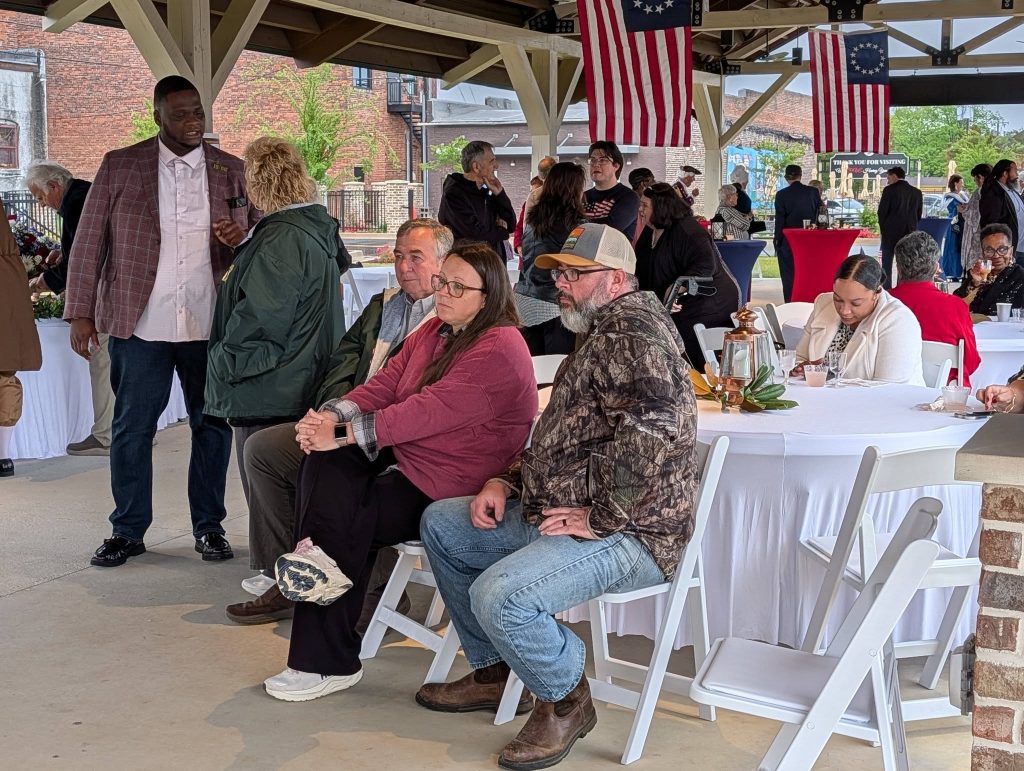 People gathered at an outdoor event under a pavilion with American flags, sitting and standing around tables covered with white cloths, engaged in conversation.