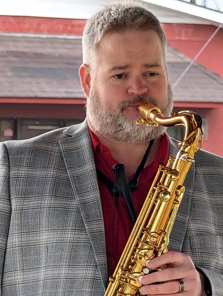 A man with a beard in a plaid jacket plays a gold saxophone outdoors, focused on the music.