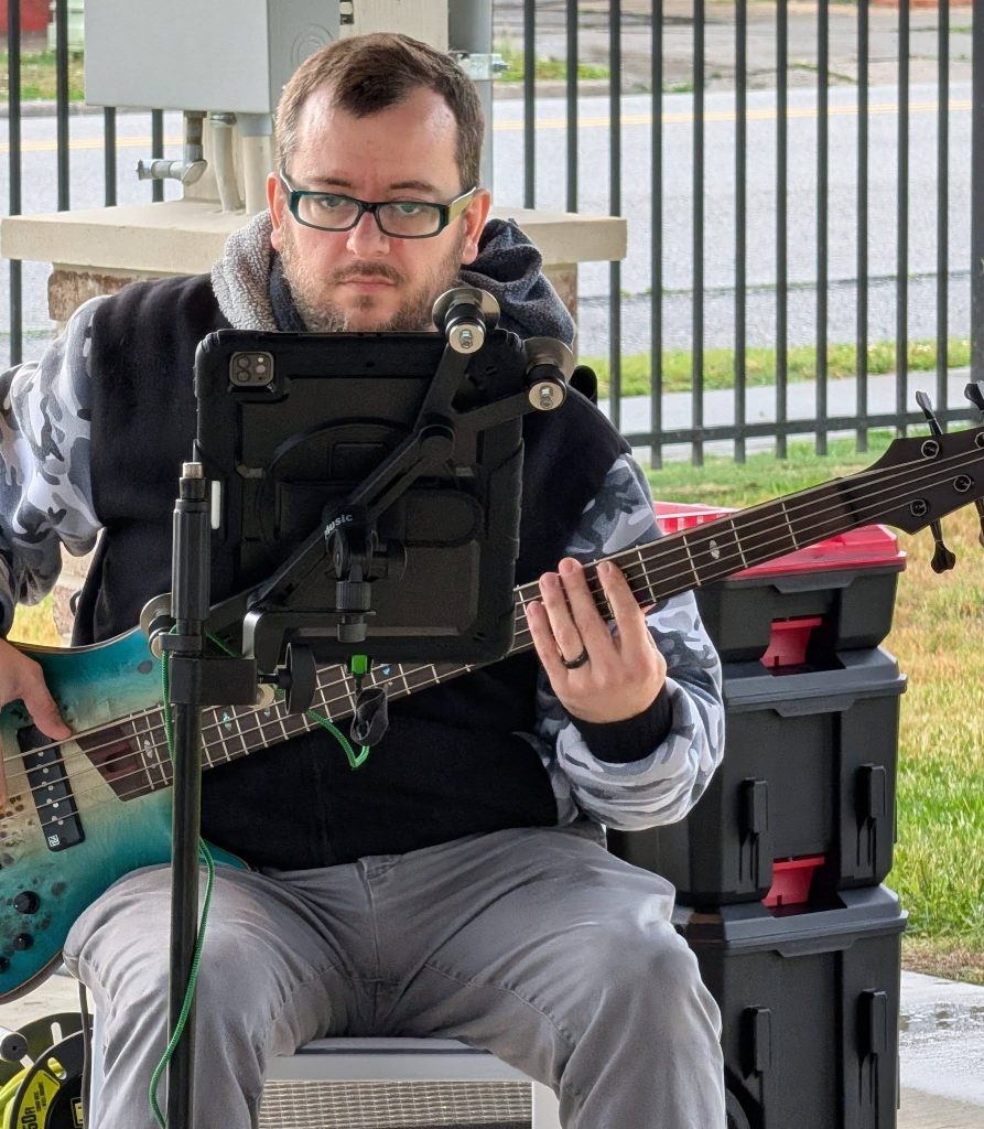 Man with glasses playing a blue electric bass guitar, seated outdoors, using a tablet mounted on a stand, with storage boxes stacked behind him.