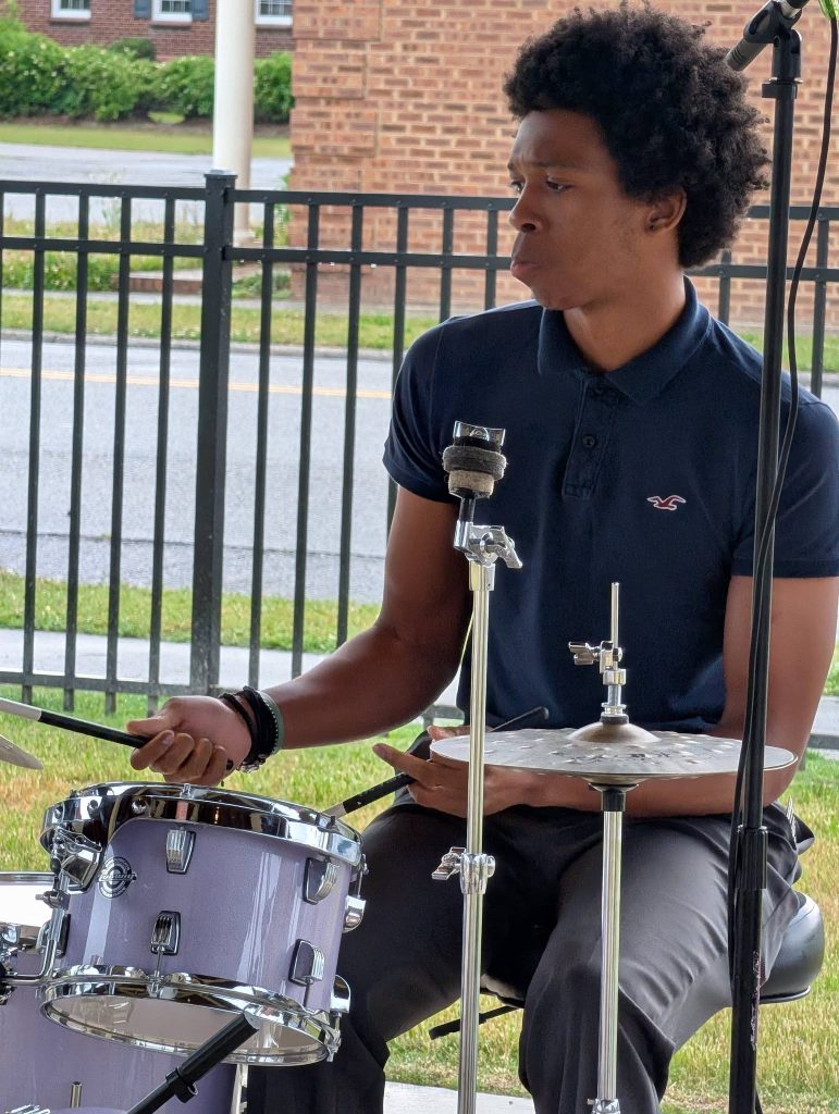 A person in a navy polo shirt plays a drum set outdoors, with a fence and brick building in the background.