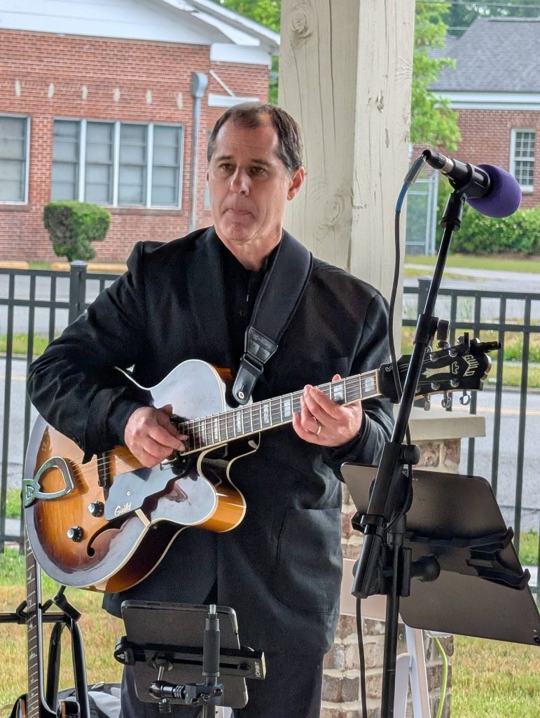 A man in a black suit plays an electric guitar near a microphone, performing outside under a covered patio.