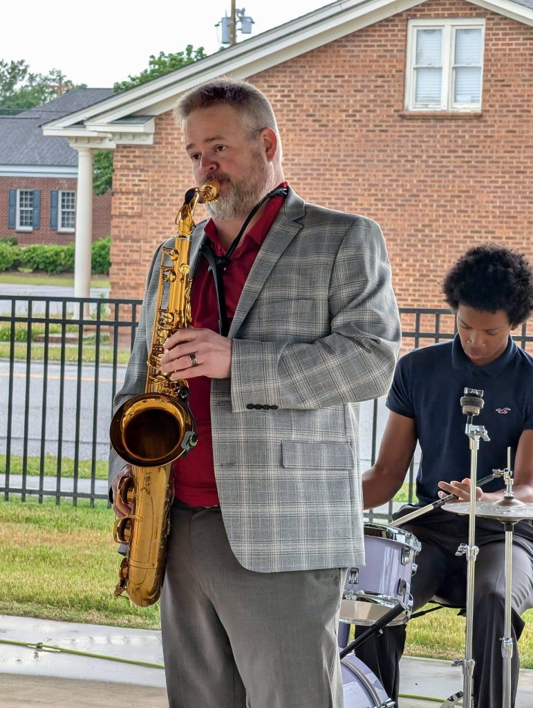 A man in a plaid blazer plays saxophone outdoors while a drummer plays in the background, with a brick building visible behind them.