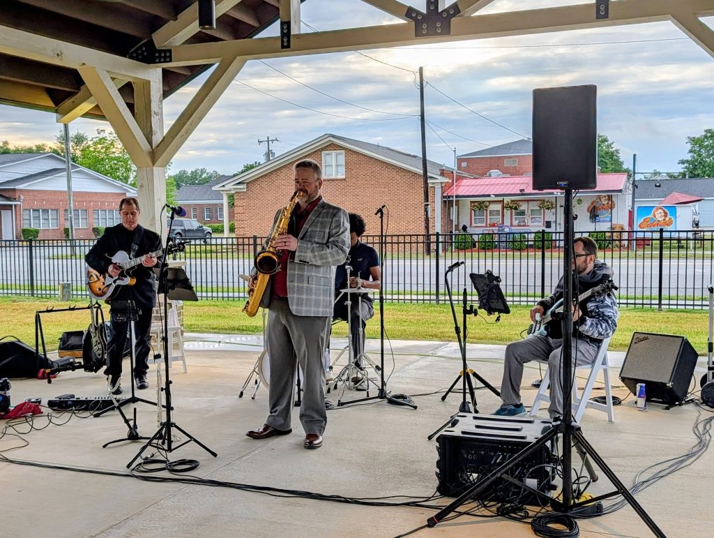 A jazz band performs under a pavilion, with musicians playing saxophone, electric guitar, bass, and drums. Outdoor setting with buildings in the background.