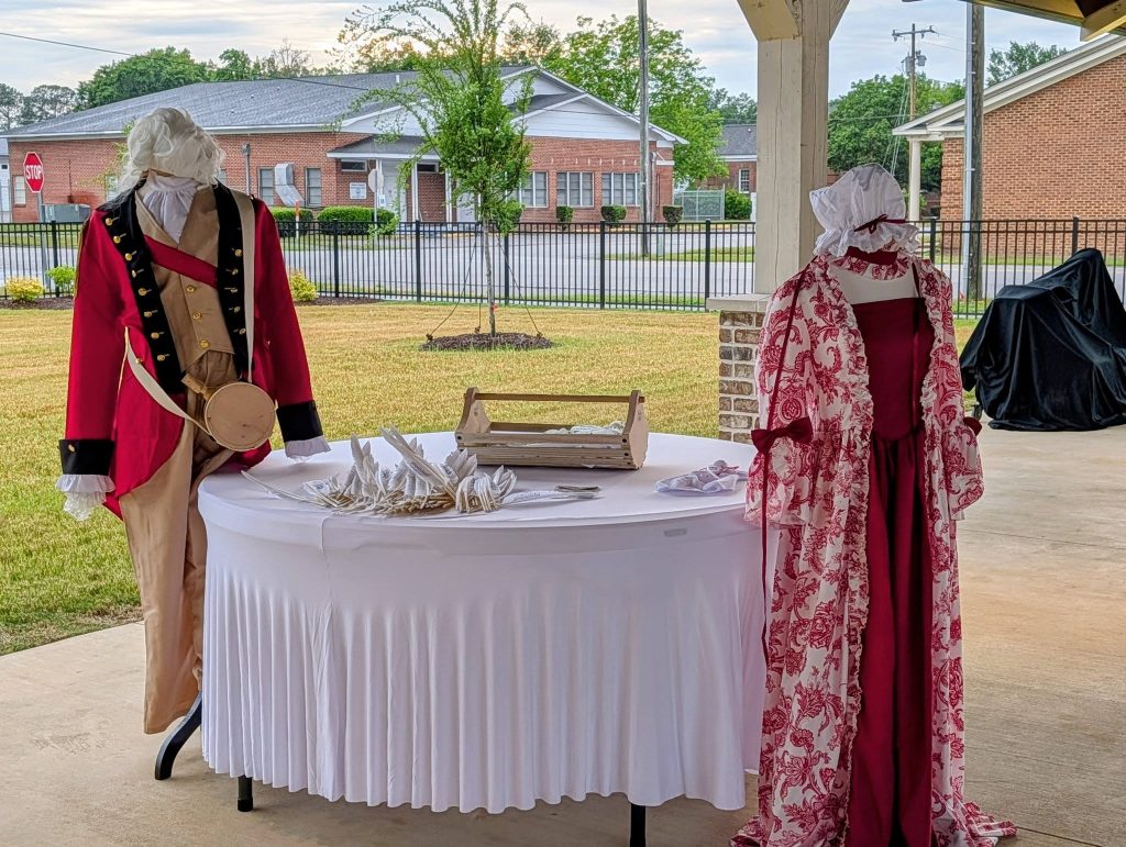 Two mannequins in historical attire stand by a round table with feathers and papers, set outdoors under a sheltered pavilion with buildings in the background.