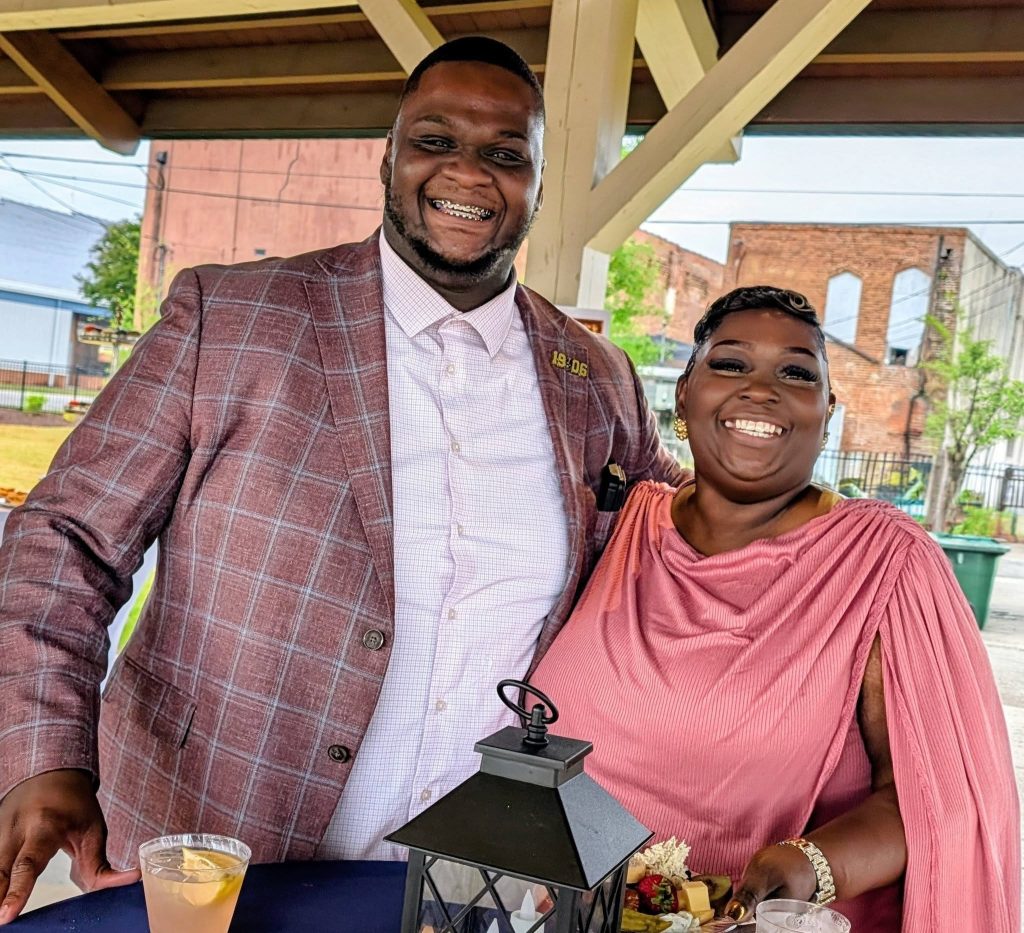 Smiling man in a plaid blazer and woman in a pink dress stand together outdoors by a table with drinks and a lantern.