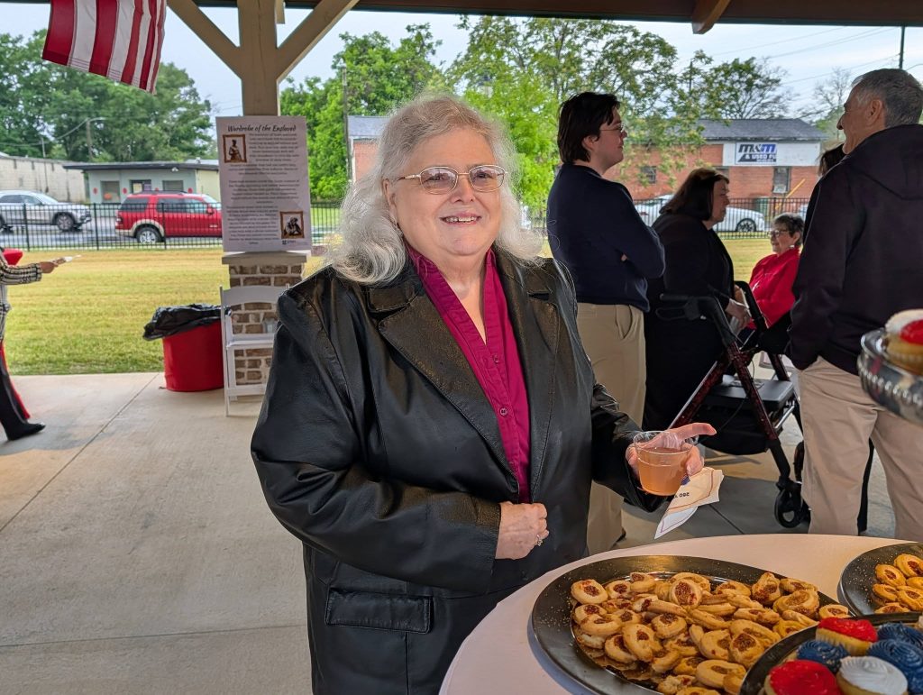 Smiling elderly woman with gray hair and glasses stands at a table with pastries, holding a drink, at an outdoor social gathering under a pavilion.