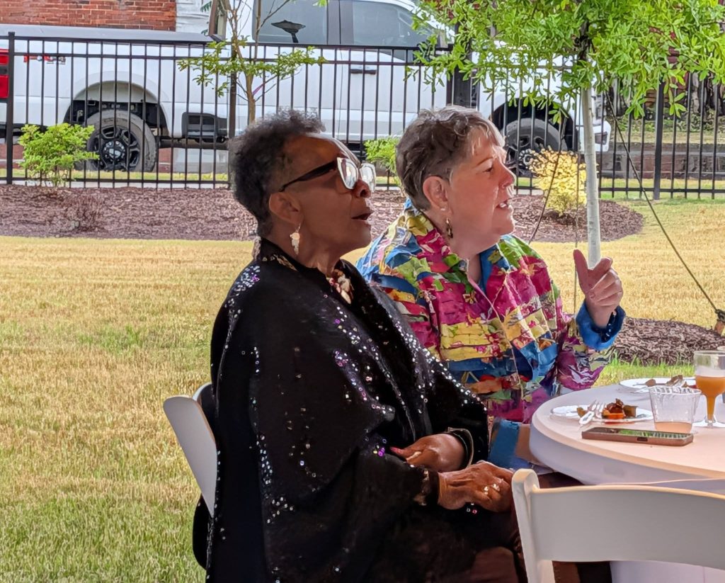 Two women sitting at a table outdoors, one in a sequined shawl and the other in a colorful jacket, engaged in conversation at an event.