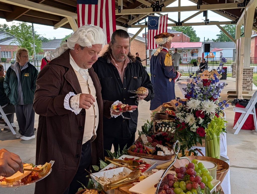People in colonial costumes at a food table with fruit, flowers, and American flags, under a pavilion during an outdoor event.