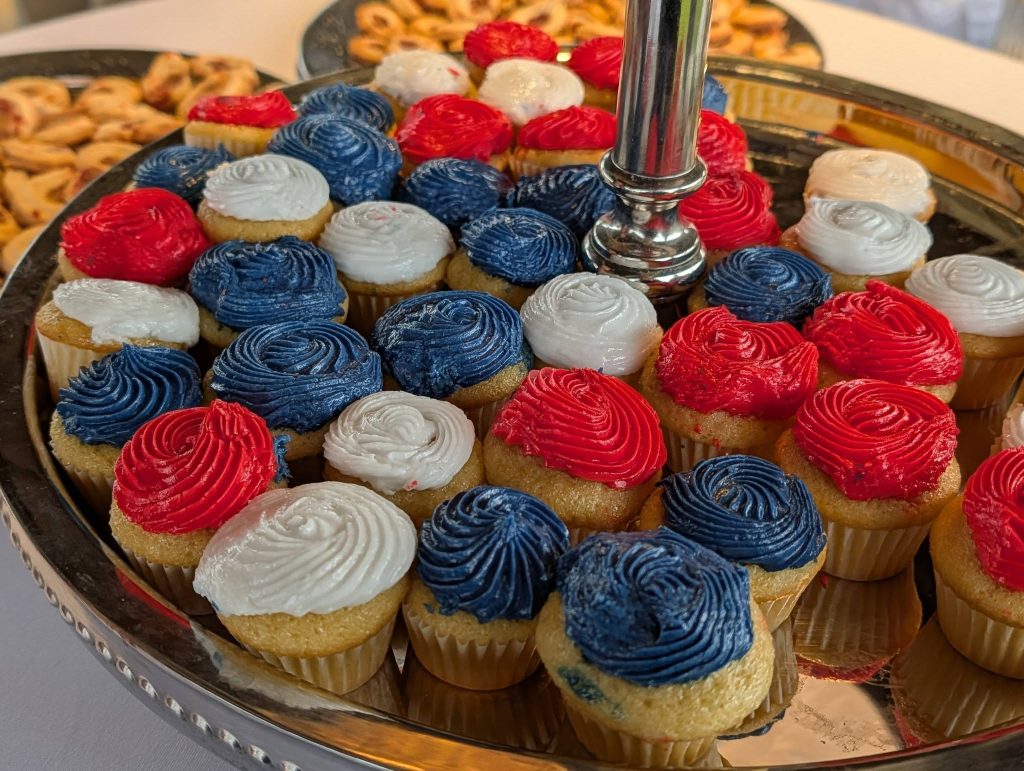 Mini cupcakes with red, white, and blue frosting arranged on a silver tray, resembling the American flag, with other desserts visible in the background.