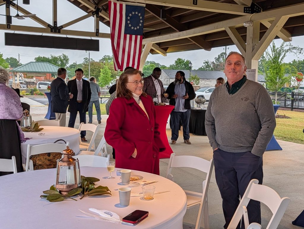 People gathered at an outdoor event under a pavilion, chatting around tables with drinks and decorations. An American flag hangs above.