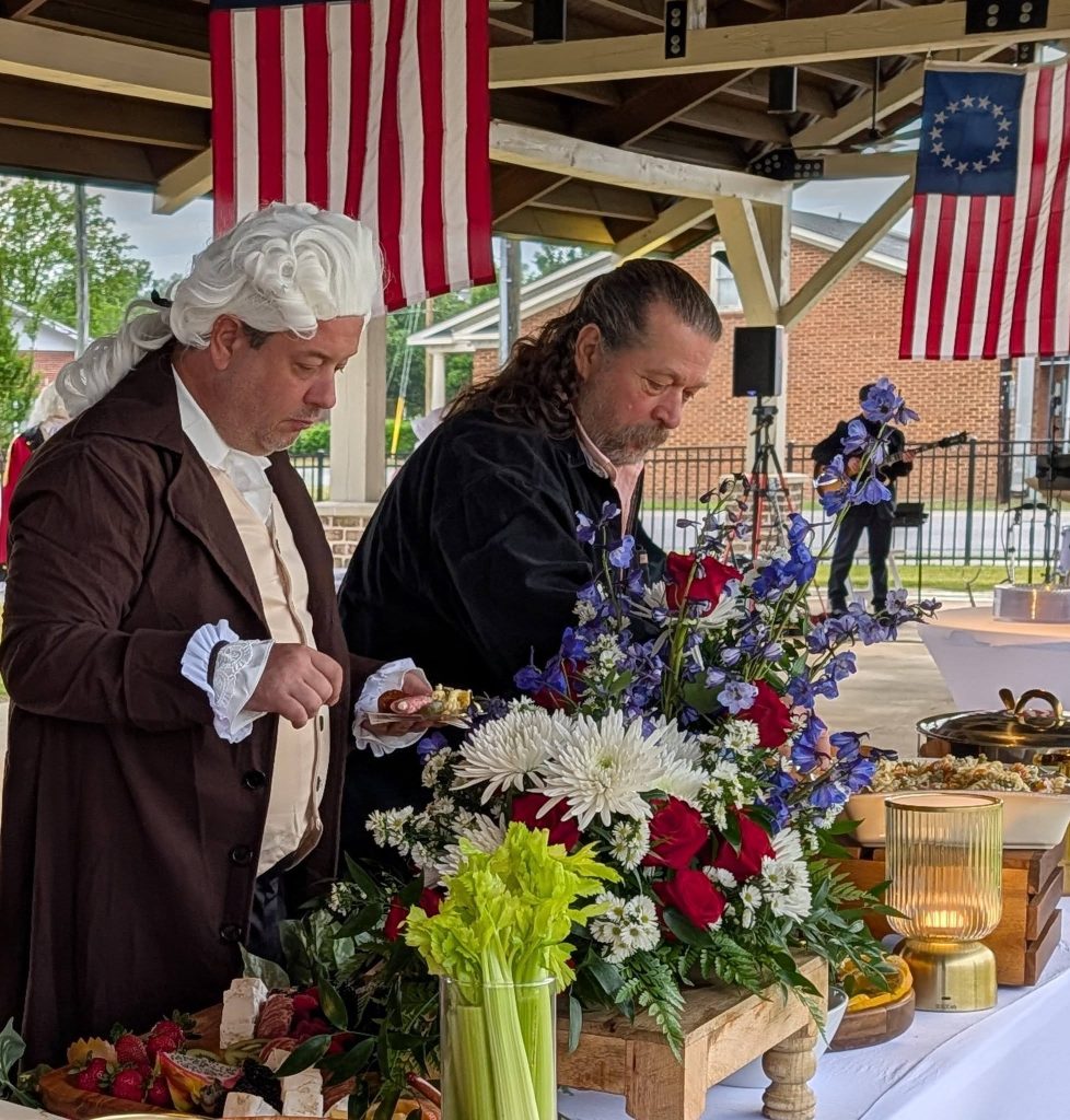 Two men in historical costumes serve themselves food at a decorated table with flowers, under American flags in an outdoor setting.