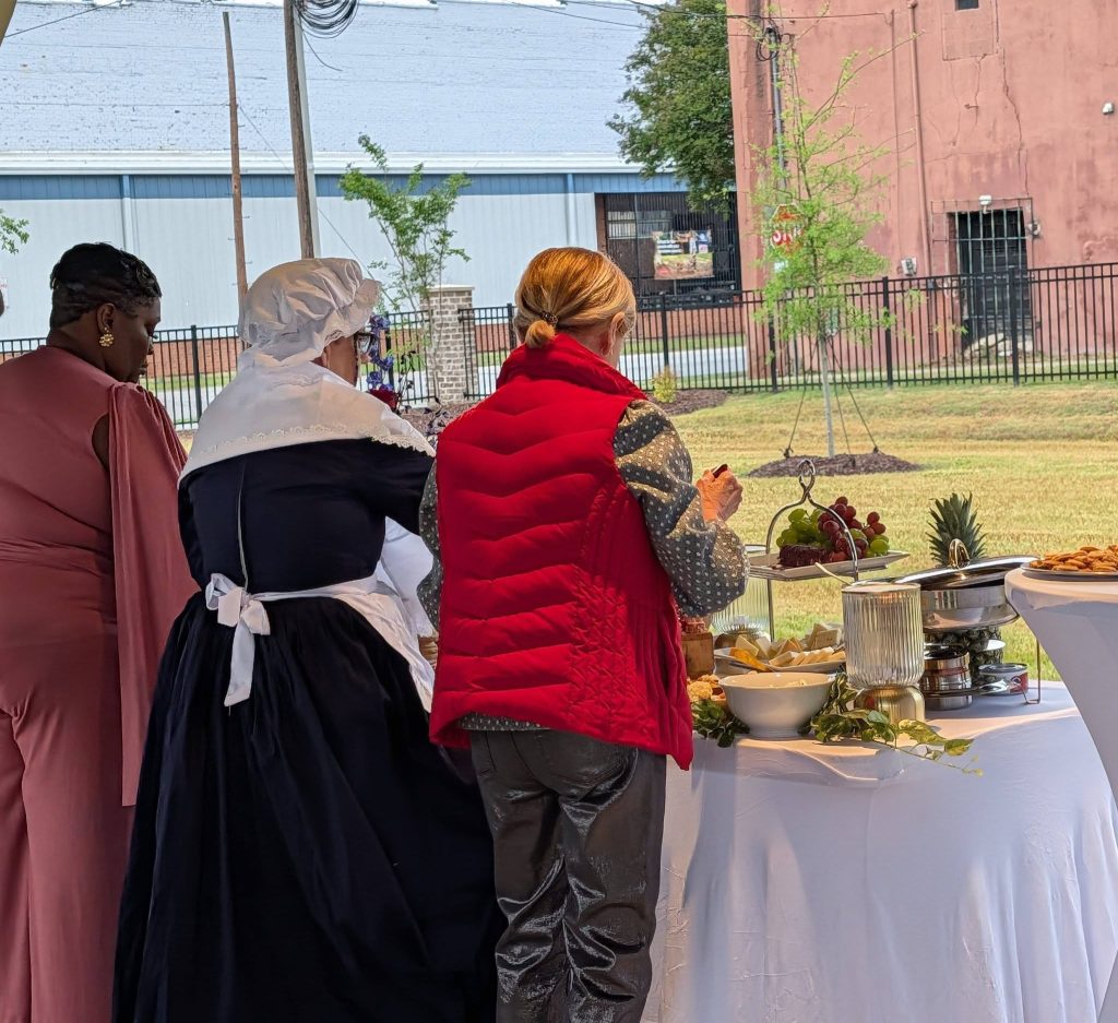 Three women stand at a buffet table outdoors, selecting food. One is dressed in historical attire, another in a red vest, and the third in a pink dress.