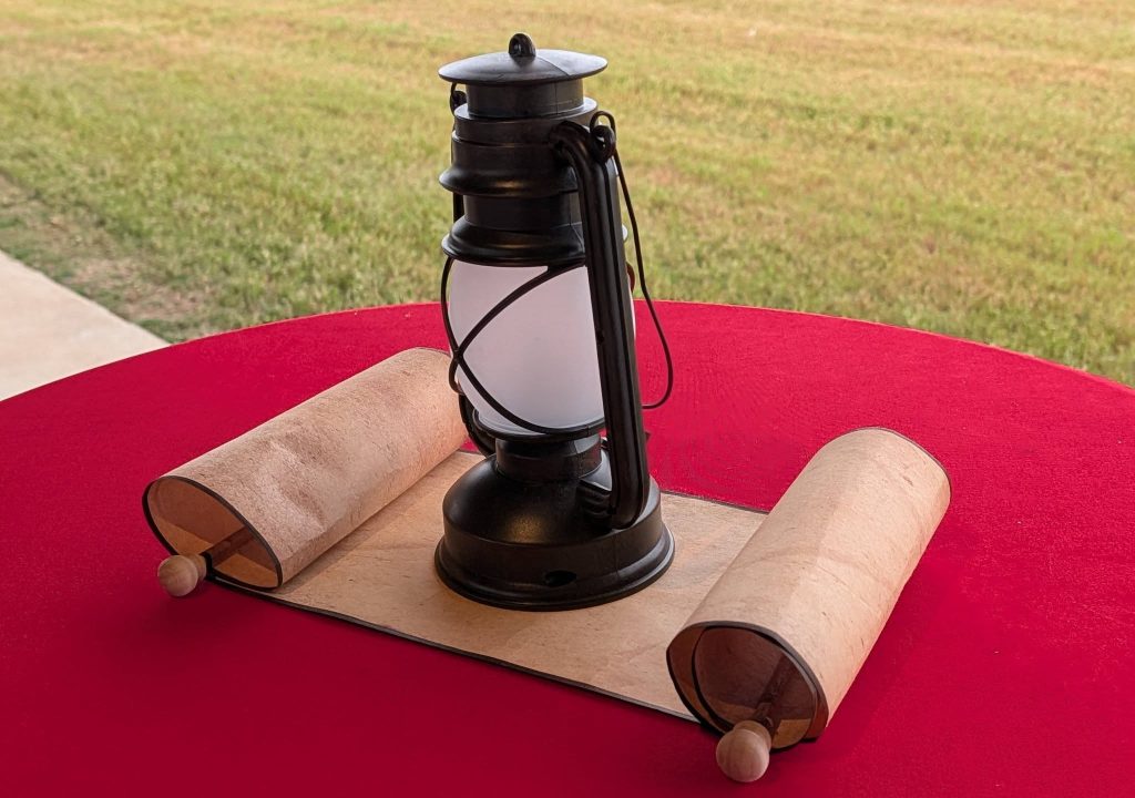 A vintage lantern and a rolled parchment sit on a red tablecloth, with grass visible in the background.
