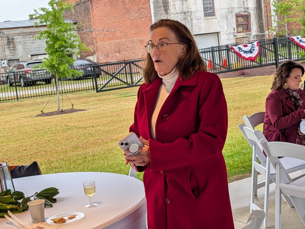 Woman in a red coat holding a smartphone stands near a table with food and drink at an outdoor event, another woman sits in the background.