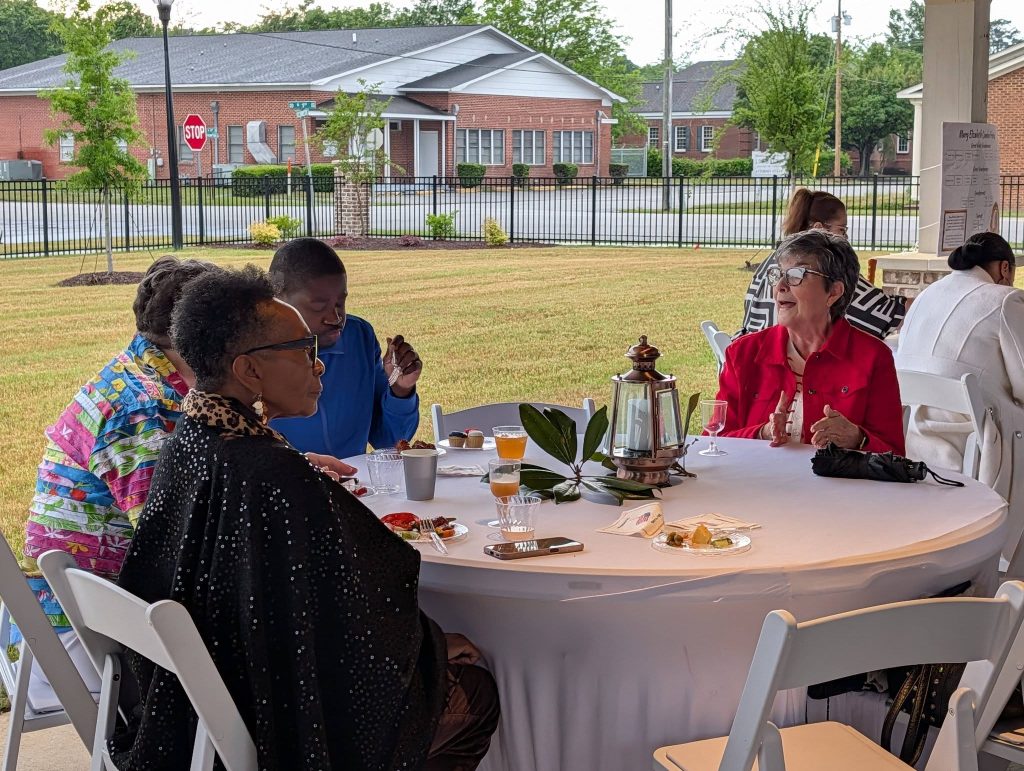 Four people sit around a round table outdoors, eating and conversing, with plates of food, drinks, and a decorative lantern on the table.