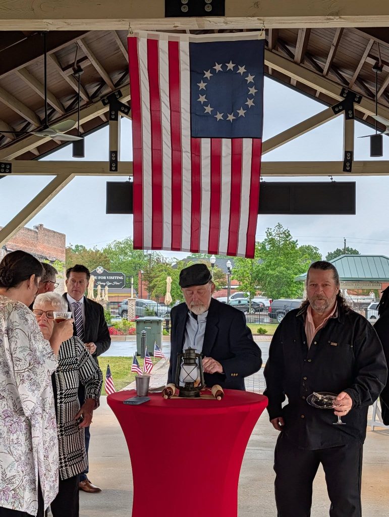 People gathered under a pavilion with a historic American flag above, standing around a red table with drinks and a lantern, American flags in background.