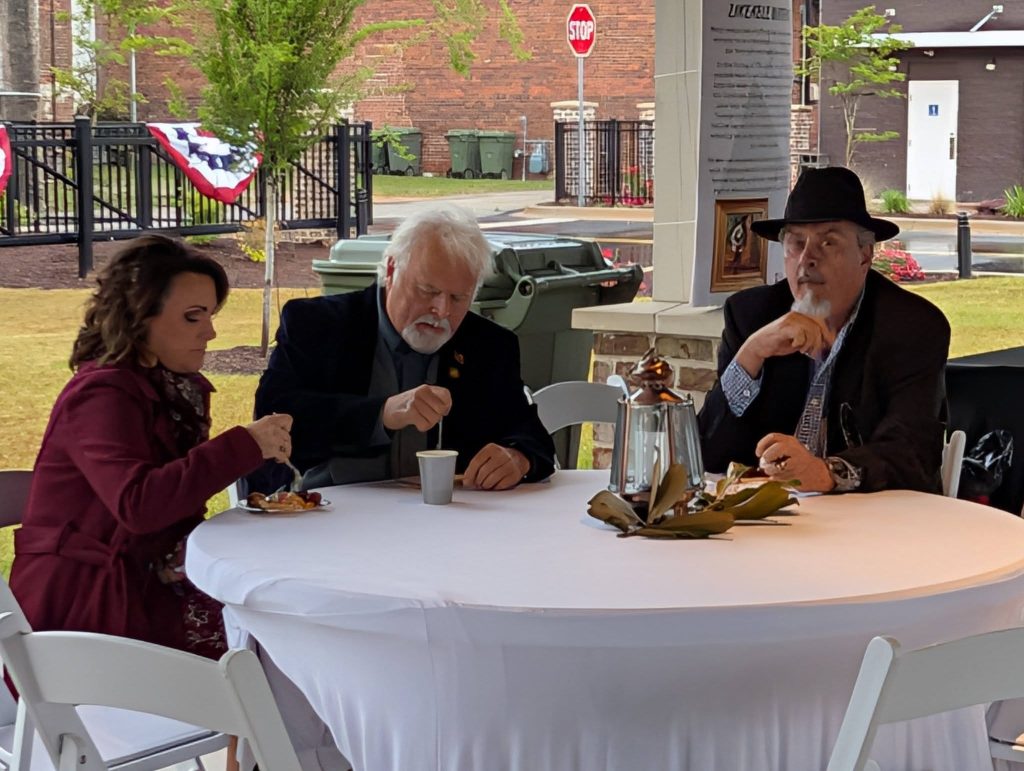 Three adults sit at a round table outdoors, eating and talking. The table is covered with a white cloth and has a decorative centerpiece and coffee pot.