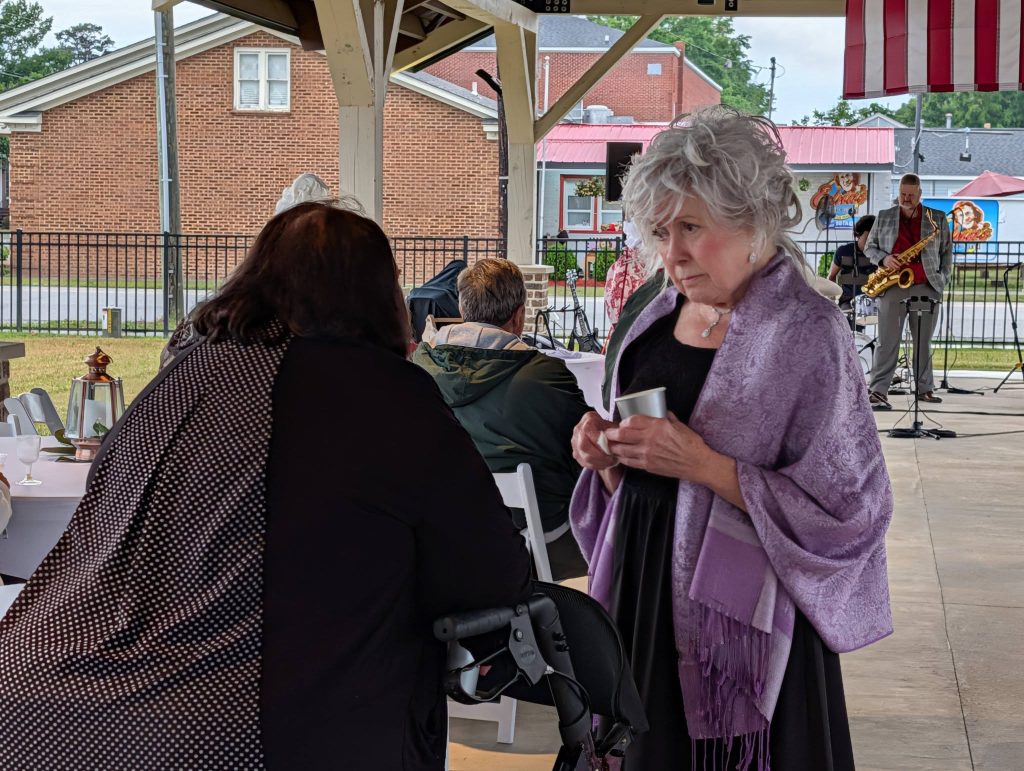Elderly woman in a lavender shawl holding a cup, talking to another woman at an outdoor gathering with live music in the background.