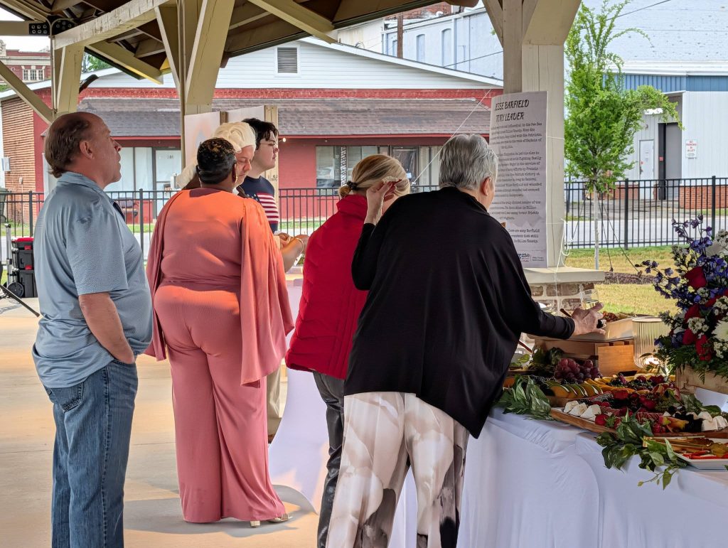 People gather under a pavilion, selecting food from a decorated table with fruit, cheese, and flowers, during a daytime outdoor event.