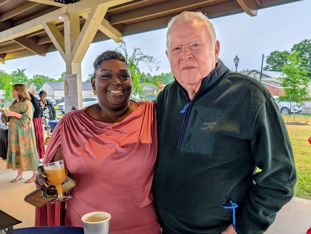 Smiling woman in a pink dress holding a drink stands next to an older man in a black jacket at an outdoor event.