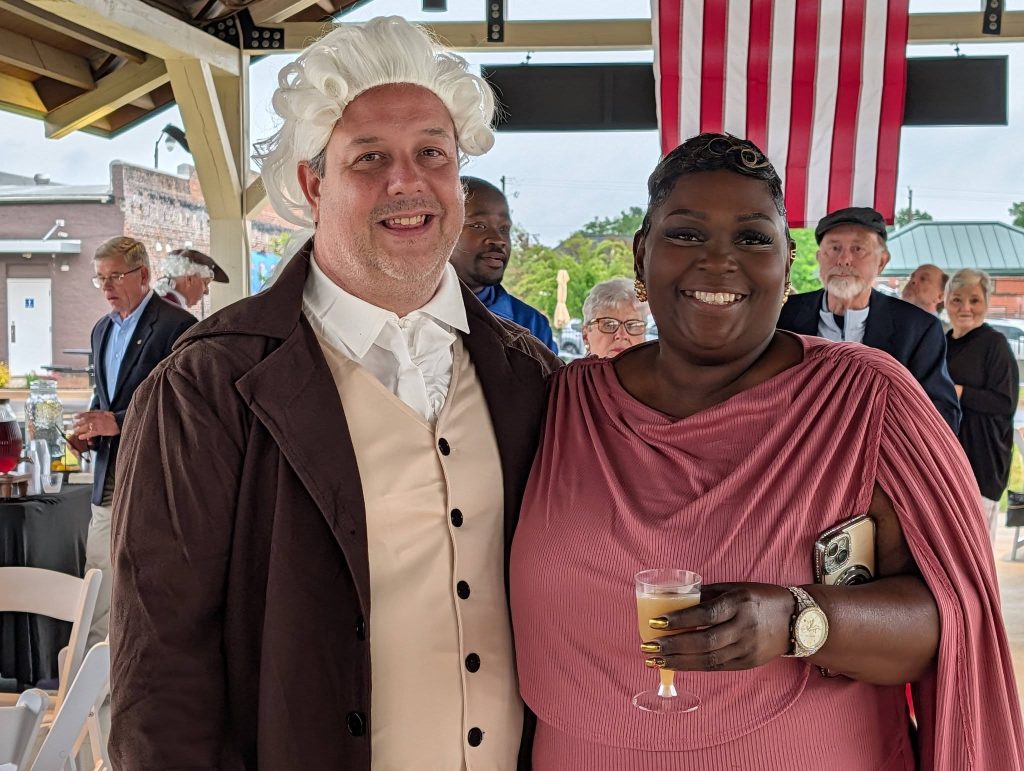 A man in historical costume and a woman in a pink dress smile at a social event with people and an American flag in the background.