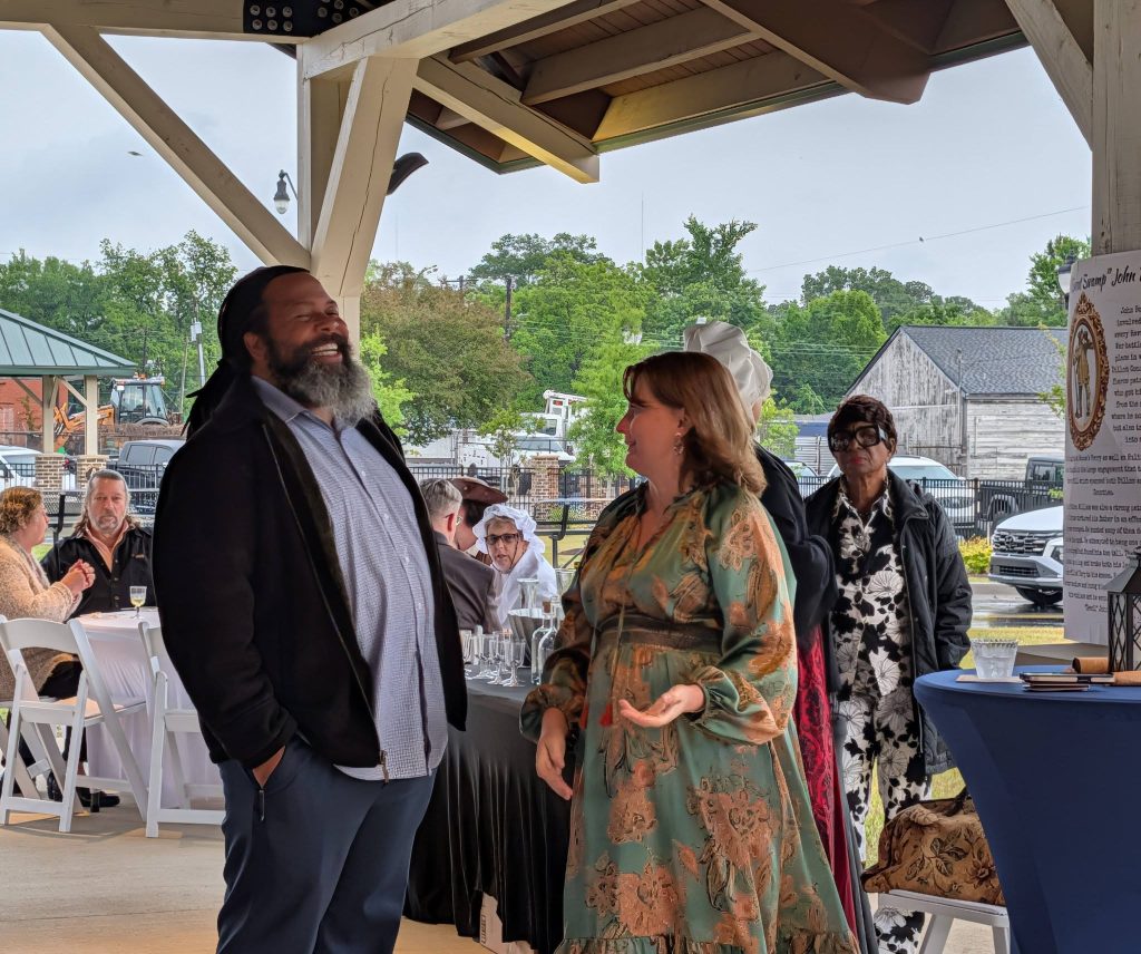 People dressed in semi-formal and period attire converse and laugh at an outdoor covered event with tables and guests in the background.