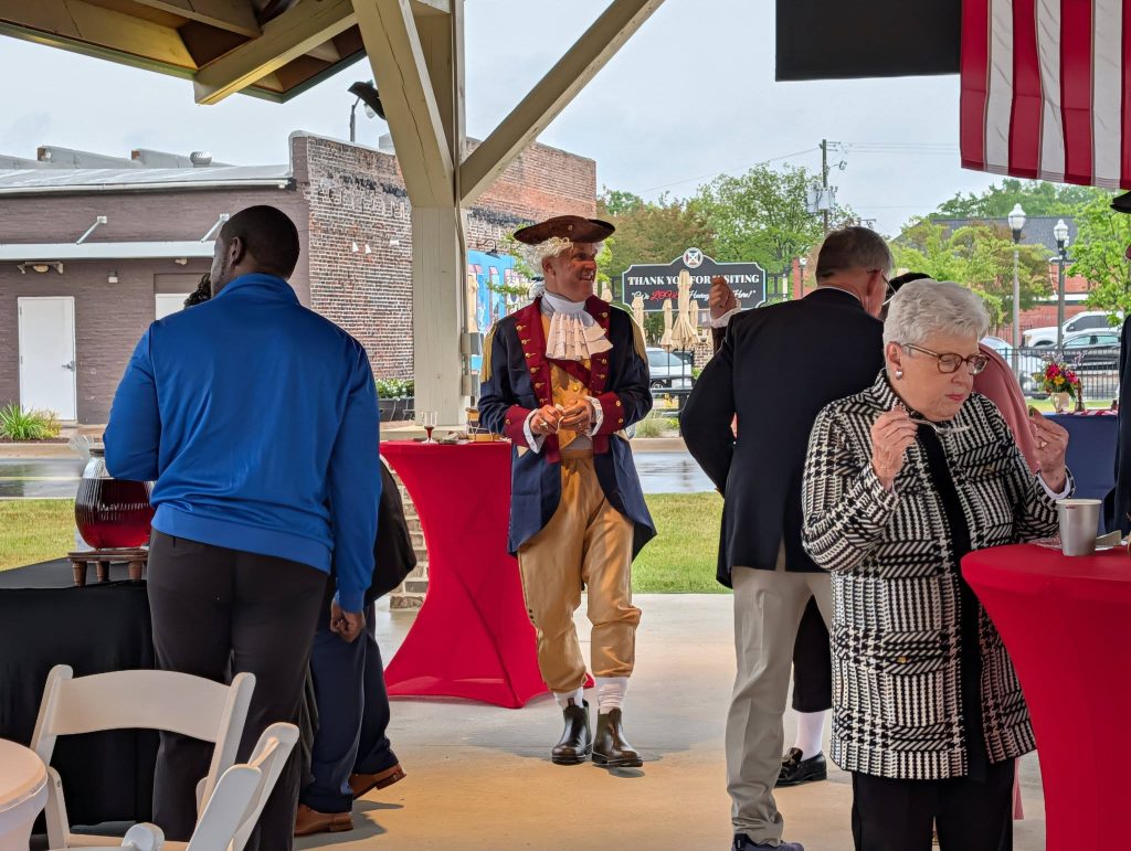 People gather at an outdoor event, one dressed in colonial attire, others in modern clothes, standing near red and black tables, under a pavilion with an American flag.