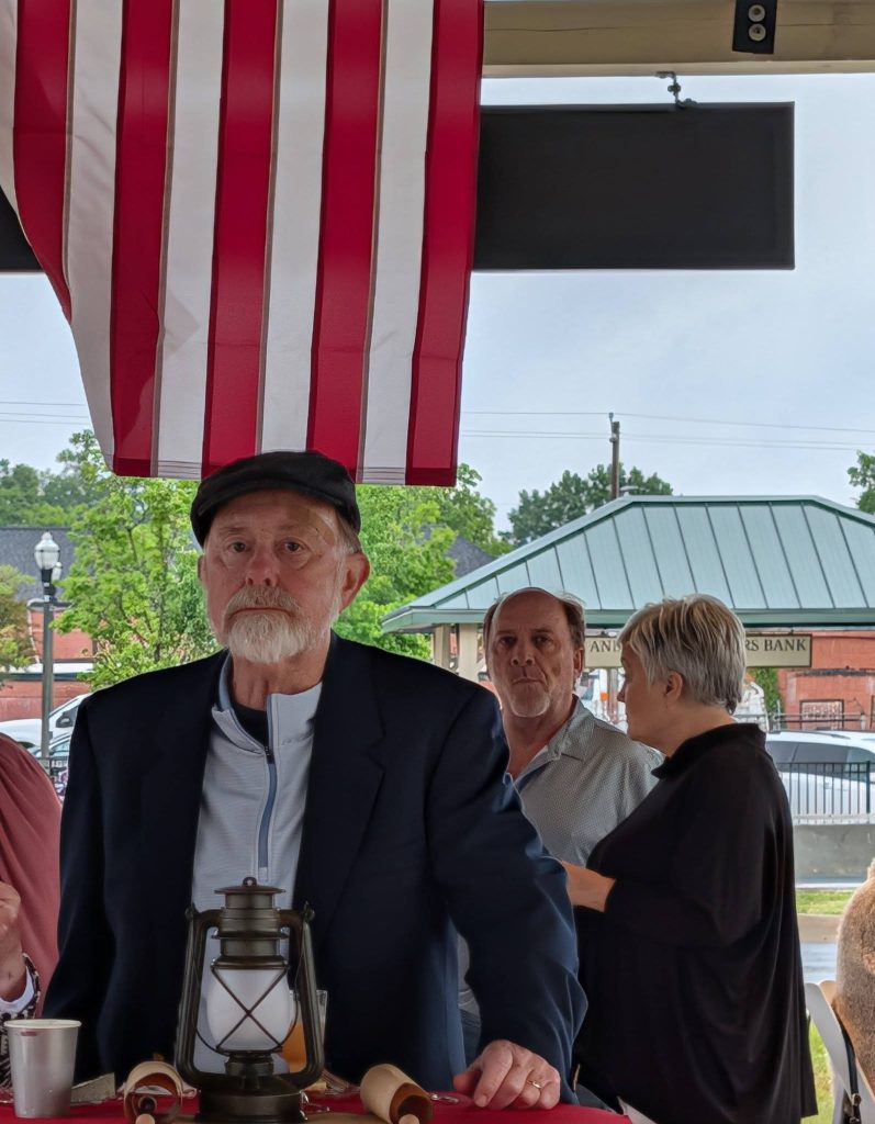 elderly man in a navy blazer stands at a table with a lantern, under an American flag, with two people conversing in the background outdoors.