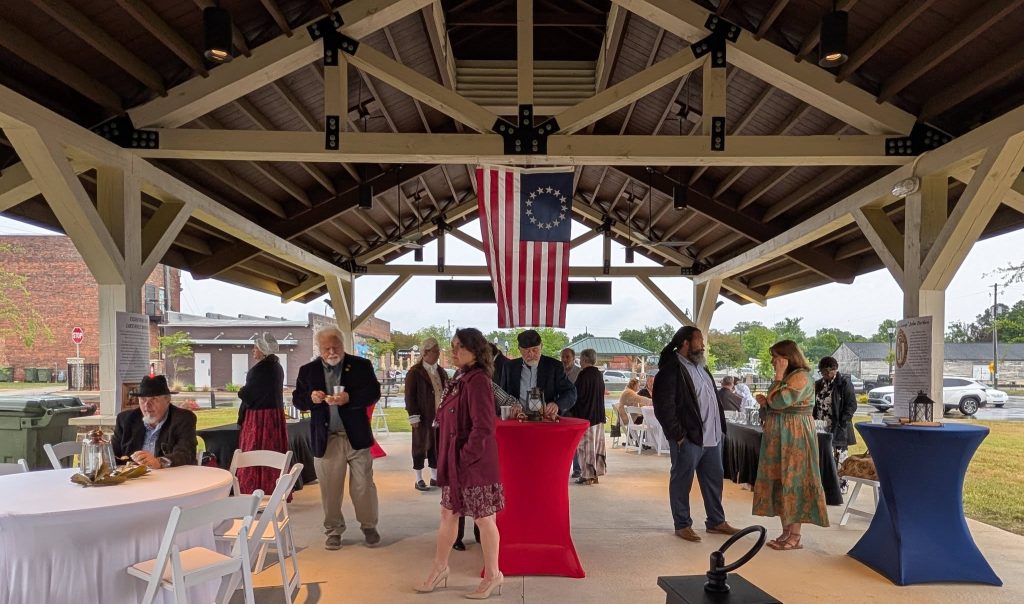 People mingling under a pavilion decorated with a historical American flag, dressed in a mix of modern and colonial-style clothing, during a daytime outdoor event.