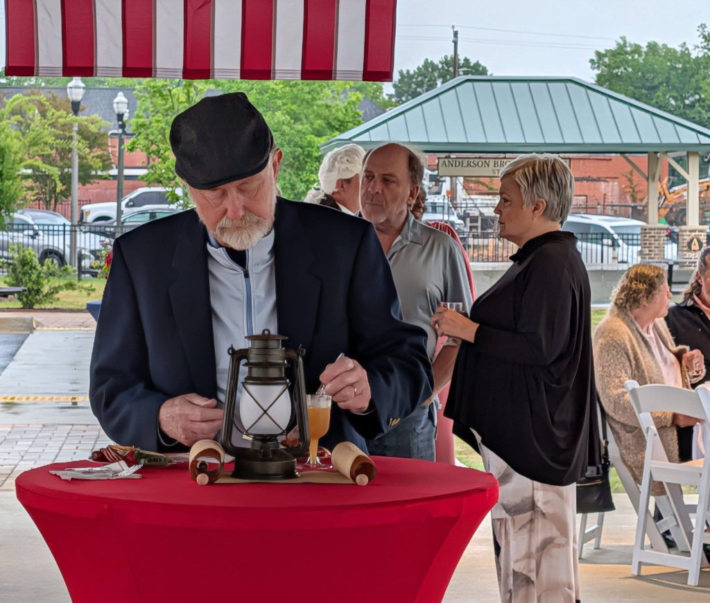 elderly man stands at a red table with a lantern, scrolls, and a drink, while other people converse in an outdoor pavilion setting.