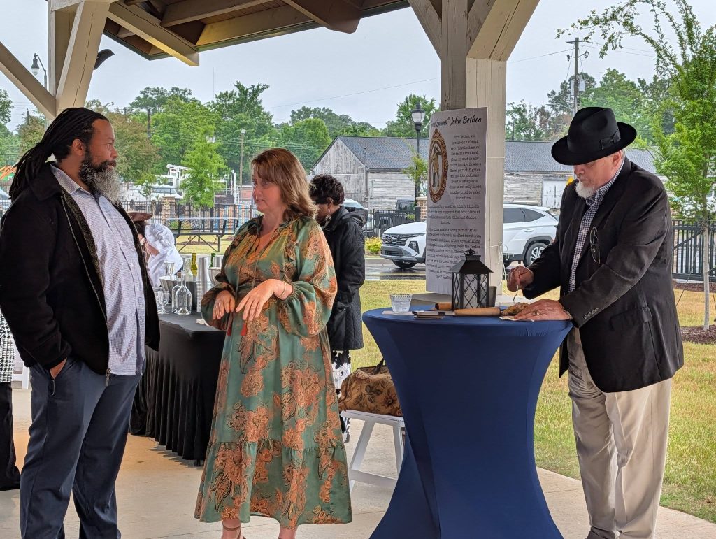 Three adults socialize at an outdoor covered event; two stand talking while one writes at a tall table with a lantern and historical display.